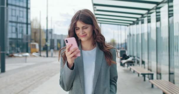 Веселая девушка тысячелетия смотрит на экран смартфона и ходит по улице. Молодая женщина-менеджер улыбается, общаясь в социальных сетях и используя телефон. На открытом воздухе . — стоковое видео