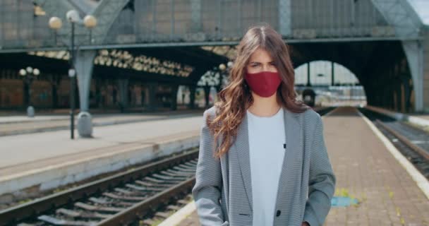 Портрет тисячолітньої жінки в багаторазовій масці для обличчя, що стоїть на залізничній станції. Вид на красиву брюнетку, яка повертає голову і дивиться на камеру. Концепція пандемії вірусу . — стокове відео