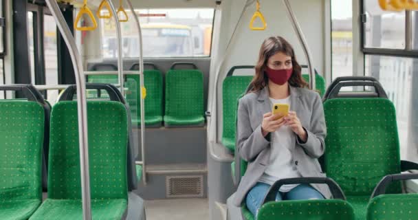 Молода брюнетка у бавовняній масці сидить у порожньому автобусі та переглядає Інтернет. Міленіаліті-жінка використовує смартфон, прокрутку та сенсорний екран. Концепція вірусу та пандемії . — стокове відео