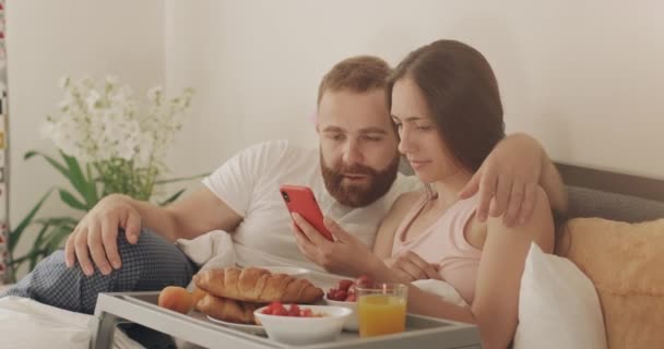 ベッドの上で横になっている間、若い男と女は電話画面を見て良いニュースを持っています。ベッドルームで朝食中に驚きの顔と木材を使用して幸せなカップル. — ストック動画