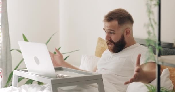 Vista lateral do cara adulto fechando seu computador portátil, gesticulando e olhando para a janela. Homem barbudo na casa dos 30 parece irritado e bebendo suco enquanto estava sentado na cama. Conceito de más notícias . — Vídeo de Stock