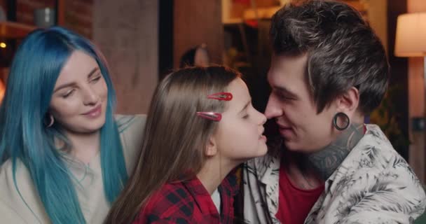 Close up of happy modern family having fun at home together. Young dad with tattoos and ear tunnels rubbing noses with his daughter while sitting near smiling mom. Concept of leisure. — Stock Video