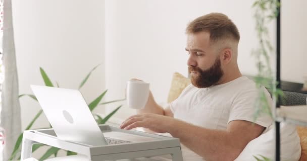 Вид сбоку приятного на вид парня, сидящего утром на кровати и улыбающегося. Бородатый мужчина-фрилансер 30-ти лет, работающий из дома и пользующийся ноутбуком за чашкой кофе . — стоковое видео