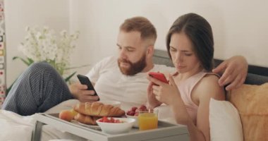 Genç çift sabah yatakta yatarken akıllı telefonlarını kullanıyor. Kadın ve erkek yatak odasında kahvaltı ederken telefon ekranlarına bakıyor. İlişki ve internet bağımlılığı kavramı.