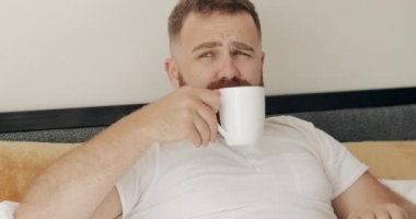Yatakta uzanıp eğlenirken kahve içen neşeli adamın portresi. 30 'lu yaşlarda sakallı yakışıklı bir adam sabahları gülümserken ve dinlenirken sıcak içkisini yudumlarken..