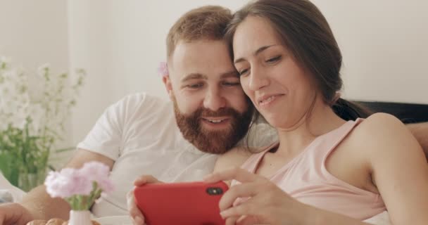 Close-up uitzicht van glimlachend jong paar met behulp van smartphone, terwijl het horizontaal houden.Gelukkig man en vrouw praten en kijken naar het telefoonscherm tijdens het ontbijt op bed. — Stockvideo