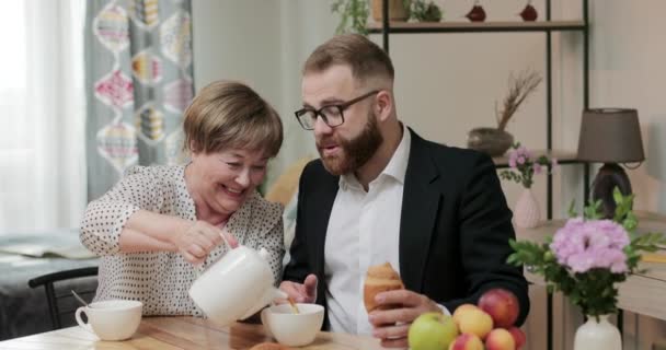 Feliz anciana sirviendo el té de su hijo en la taza y sonriendo. Joven hombre de negocios comiendo croissant y comunicándose mientras visita a su madre en casa. Concepto de amor y cuidado . — Vídeos de Stock
