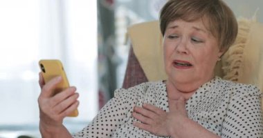 Yetişkin emeklilerin modern cep telefonlarını kullanırken kötü haberler alması üzücü. Yaşlı bir kadın oturmuş telefon ekranına bakarken kafasını sallıyor ve üzülüyor..