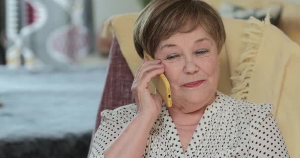 60 'lı yaşlarda neşeli yaşlı bir bayanın modern cep telefonuyla konuşması ve gülümsemesi ön planda. Mutlu bir emekli kadın, oturma odasında sandalyesinde otururken arkadaşıyla sohbet ediyor.. — Stok video