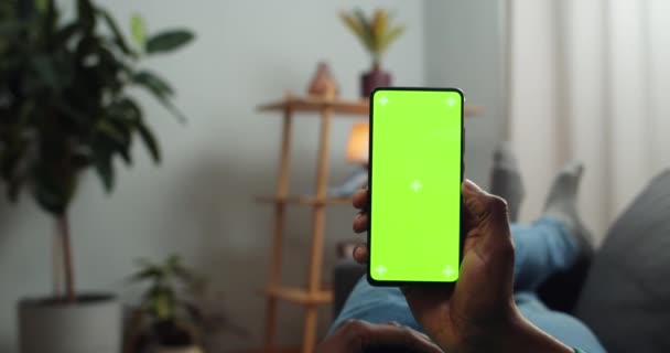 Vista de la cámara del hombre africano sosteniendo un teléfono inteligente moderno con pantalla verde mientras está acostado en el sofá. Concepto de croma key, maqueta y tecnología. Acogedor fondo casero . — Vídeos de Stock