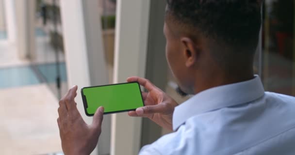 Overschouderbeeld van een Afro-Amerikaanse man met een telefoon in horizontale landschapsmodus. Kerel kijken en aanraken van groene smartphone scherm. Concept van chroma sleutel. Moderne bouwachtergrond. — Stockvideo