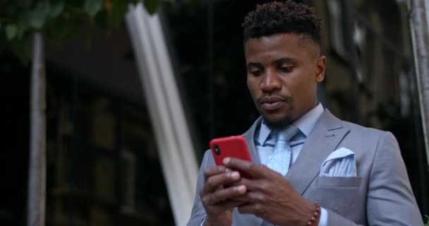 Hombre afroamericano serio en traje clásico a medida y corbata navegando por Internet y tocando la pantalla del teléfono. Chico exitoso en ropa elegante usando teléfono inteligente mientras está parado en la calle . — Vídeos de Stock