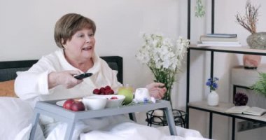 Mutlu yaşlı kadın yemek dolu Trey 'le yatakta otururken televizyonu değiştiriyor. Gülümseyen olgun kadın kahvaltı yaparken televizyon ekranına bakıyor ve günaydın diyor. Rahatlama kavramı.