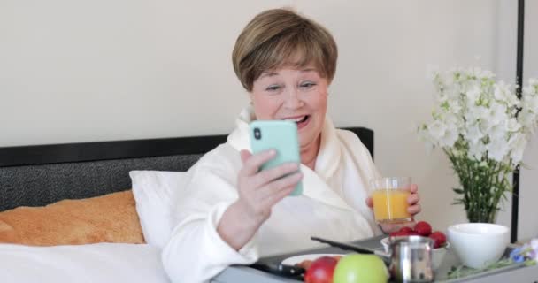 Alegre anciana mirando la pantalla de su teléfono inteligente moderno y riendo. Mujer anciana de buen humor sosteniendo un vaso de jugo y usando el móvil durante el desayuno en la cama . — Vídeos de Stock