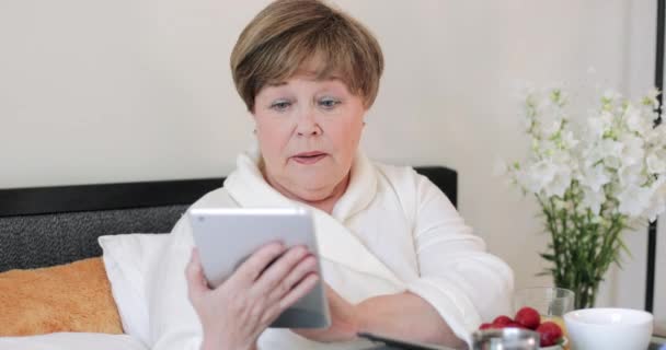 Alte Dame mit ernstem Gesichtsausdruck, die Hausrat trägt und Tabletten nimmt. Ältere Rentnerin sitzt auf dem Bett und frühstückt, während sie auf den Bildschirm ihres modernen Geräts blickt. — Stockvideo