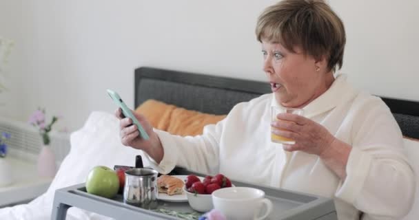 彼女の近代的な携帯電話を使用している間に驚くべき顔の表情を作る老婦人のサイドビュー。衝撃的なニュースを取得しながら、朝食やジュースを飲んでいる高齢女性. — ストック動画