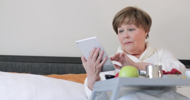 Vista inferiore di grave signora anziana digitando sullo schermo del tablet durante la colazione. Bella donna anziana utilizzando il suo gadget moderno mentre seduto sul letto con trey pieno di cibo sulle gambe . — Video Stock