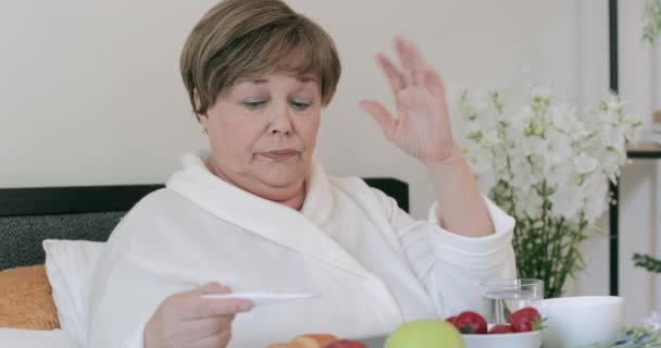 Stará žena chytí rýmu a drží teploměr při snídani v posteli. Žena v důchodu v šedesáti se cítí špatně, zatímco sedí s Treyem plným jídla. Koncepce lidí a zdravotní problémy. — Stock video
