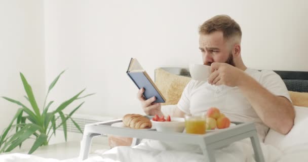 30 'lu yaşların ortasında, kahve içerken kitap okuyan entelektüel bir adam. İyi görünümlü sakallı adam yatakta otururken kahvaltı yapıyor ve kitap sayfasını çeviriyor. Boş zaman kavramı ve günaydın.. — Stok video