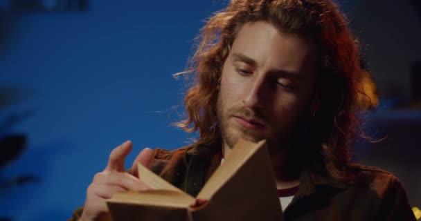 Roman okurken sayfayı çeviren yakışıklı genç adamın yakın görüntüsü. Uzun saçlı ve sakallı milenyum çocuğu akşamları boş vakit geçirirken kitap okuyor. Ev arkaplanı. — Stok video