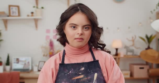 Close up of adorable teen with down syndrome wearing in artists apron and holding paintbrush. Portrait of pretty girl looking and posing to camera in her room. Concept of sunny children. — Stock Video