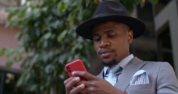 Zamknij widok poważnego biznesmena w kapeluszu i garnitur wpisując wiadomość i patrząc na ekranie telefonu. Poważny facet w stylowych ubraniach za pomocą smartfona do komunikacji na ulicy. — Wideo stockowe