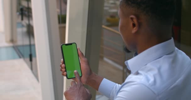Vista sobre el hombro del hombre afroamericano sosteniendo el teléfono inteligente y tocando la pantalla verde mientras hace un gesto de sí. Clave de croma, burla.Concepto de éxito. Edificio de oficinas moderno fondo . — Vídeo de stock