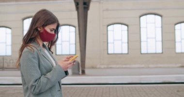 Bin yıllık esmer bir kızın akıllı telefon kullanıp yürümesinin yan görüntüsü. Boş tren istasyonunda el yapımı maske takmış genç bir kadının ekran görüntüsü kayıyor ve dokunuyor.