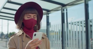 Akıllı telefon kullanan ve yürüyen koruyucu maskeli bir kızın yakın plan fotoğrafı. Milenyum kızı internette gezinirken telefon ekranına bakıyor. Salgın ve koronavirüs kavramı.