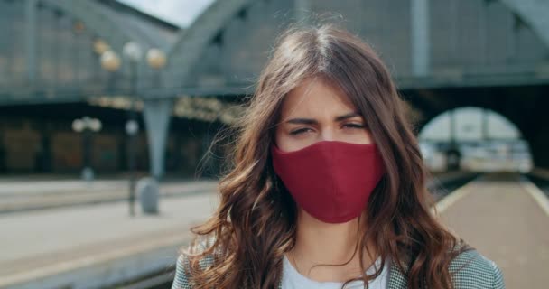 Close up view of brunette woman in cotton protective face mask standing at railway station. Portrait of serious young girl looking to camera. Blurred background. Concept of virus pandemic. — Stock Video