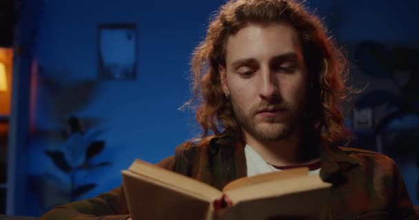 Närbild av koncentrerad ung kille vända blad medan du läser litteratur. Smart man med långt hår och skägg enjoing intressant bok samtidigt tillbringa fritid hemma. — Stockvideo