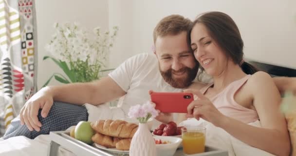 Happy guy and girl watching funny video and laughing in bed. Young romantic family having breakfast and talking while looking at smartphone screen in early morning. Concept of leisure. — Stock Video