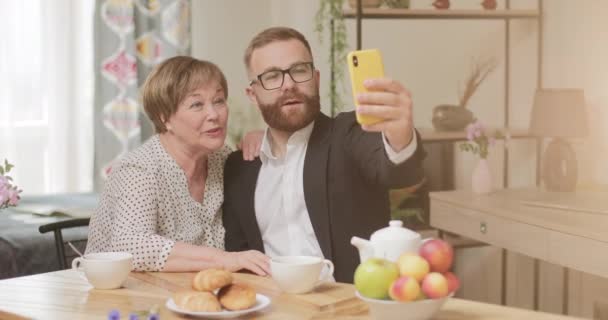 Vooraanzicht van gelukkige zoon en zijn volwassen goed uitziende moeder die plezier hebben en selfie nemen. Man in pak maken foto met oudere gepensioneerde vrouw op smartphone thuis. — Stockvideo