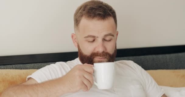 Close-up vista do jovem alegre inalando aroma enquanto sentado na cama de manhã. Happy barbudo cara na década de 30 cheirando café e sorrindo para a câmera, tendo bom humor . — Vídeo de Stock