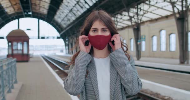 Vista de cerca de la chica atractiva del milenio quitándose la máscara de algodón mientras está de pie en la plataforma de la estación de tren. Joven mujer feliz respirando profundamente y sonriendo mientras mira a la cámara  . — Vídeo de stock