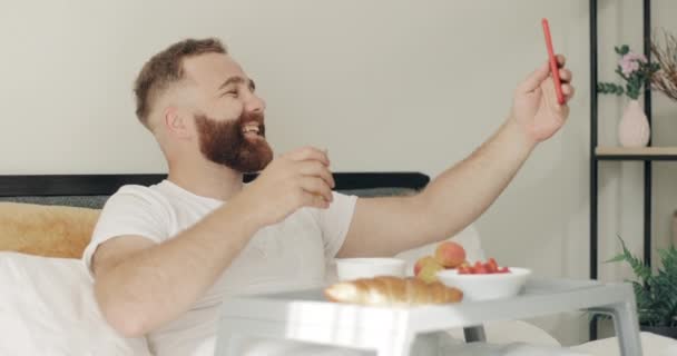 Вид сбоку улыбающегося парня, который ведет видеозвонок, сидя и завтракая в постели. Взрослый мужчина разговаривает и смеется, используя фронтальную камеру смартфона. Концепция коммуникации . — стоковое видео