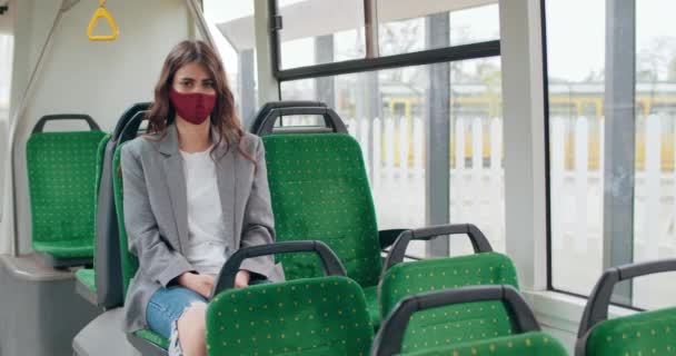 Portret dziewczyny w bawełnianej masce twarzy siedzącej w pustym autobusie i patrzącej w kamerę. Tysiącletnia brunetka w luźnych ubraniach z ochronną maską wielokrotnego użytku. Pojęcie wirusa i pandemii. — Wideo stockowe