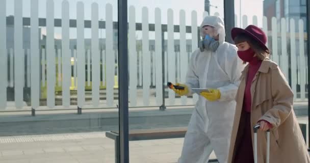 Jeune femme avec de la fièvre portant une valise tout en marchant et en parlant avec le travailleur médical en combinaison de protection et masque. Concept Symptômes de Covid-19, coronavirus et épidémie. À l'extérieur . — Video