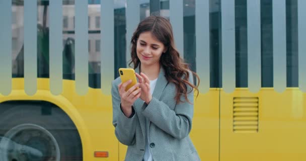 Millennial γυναίκα συγκινητικό και κοιτάζοντας οθόνη smartphone. Χαρούμενη μελαχρινή κοπέλα χρησιμοποιώντας το τηλέφωνο κατά την περιήγηση στο διαδίκτυο ή κρέμονται στα κοινωνικά μέσα μαζικής ενημέρωσης. έξω. Κίτρινο λεωφορείο στο backgrond. — Αρχείο Βίντεο