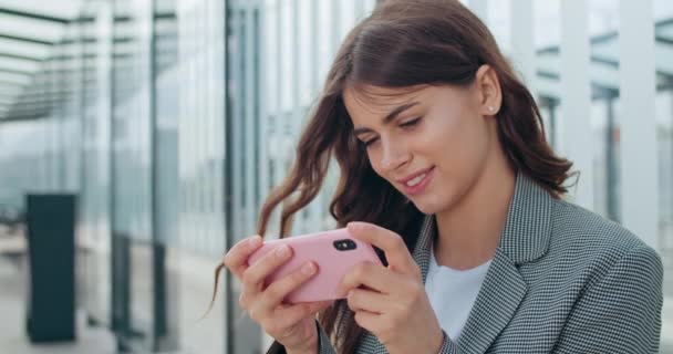 Vista de cerca de la joven attarctiva sonriendo mientras usa el teléfono inteligente cerca del edificio moderno. Chica alegre Millennial sosteniendo el teléfono en modo horizontal paisaje y pantalla táctil . — Vídeo de stock