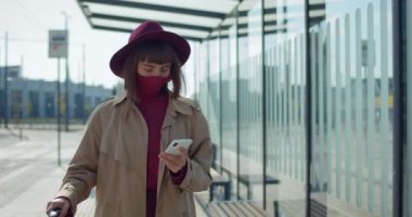 Tren istasyonunun yanında akıllı telefon kullanan koruyucu maskeli ve valizli şık bir kadın. Genç bayan gezgin yürüyor ve telefon ekranına bakıyor. Koronavirüs kavramı .