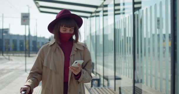 Mujer con estilo en máscara protectora y maleta con teléfono inteligente cerca de la estación de tren. Viajera joven caminando y mirando la pantalla del teléfono y a un lado. Concepto de coronavirus  . — Vídeos de Stock