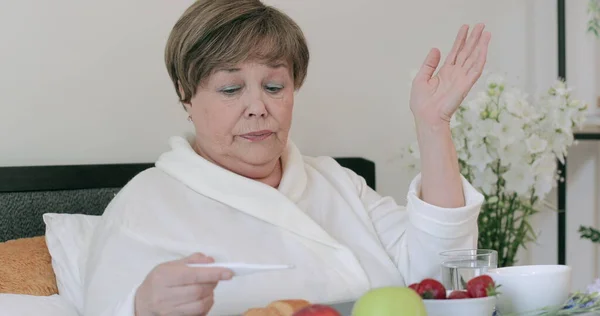老太太在床上吃早餐时感冒了,还拿着温度计.60多岁的退休女士坐在满是食物的树上感觉很糟。人与健康问题的概念. 图库图片