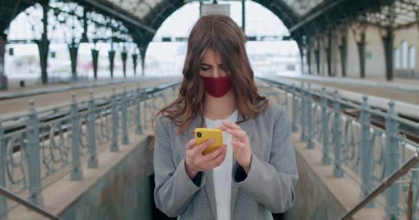 Młoda ładna dziewczyna w bawełnianej masce na twarz i dotykając ekranu telefonu. Widok plonu tysiącletniego brunetka kobieta telefon stojąc sam. Koncepcja pandemii wirusa. — Wideo stockowe