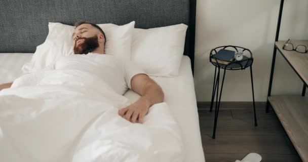 젊고 잘생긴 청년이 자고 있고 침대에 누워 있는 동안 매우 빠르게 일어나고 있습니다. 수염 이 많은 남자가 아침에 늦잠을 자는 모습을 보면서 일어나 소리를 지르고 있습니다. — 비디오