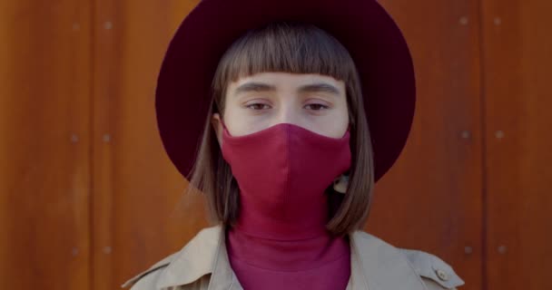 Close up view of pretty girl in protective cotton mask looking to camera and blinking. Portrait of stylish young woman in hat with reusable mask on face . Zoom in. Rust background. — Stock Video
