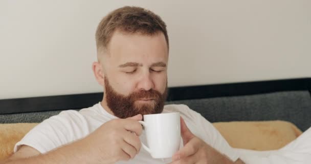 Close up view of happy adult man drinking coffee with rich aroma while sitting on bed in morning.Νεαρός γενειοφόρος άντρας στα 30 χαμογελά και απολαμβάνει το πρωί ενώ έχει καλή διάθεση. — Αρχείο Βίντεο