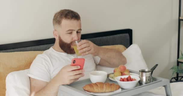 Sakallı genç adam meyve suyu içerken boğuluyor ve şok edici haberler okuyor. 30 'lu yaşların ortasında yakışıklı bir adam akıllı telefonunu kullanıp yatakta kahvaltı ederken çok şaşırmış görünüyor.. — Stok video