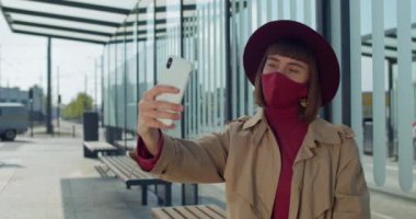 Pamuk maskeli bir kız modern cam bina yakınlarındaki sosyal ağlarda selfie çekiyor ve paylaşıyor. Akıllı telefon kamerasıyla poz veren çekici milenyum kızı. Covid 19.