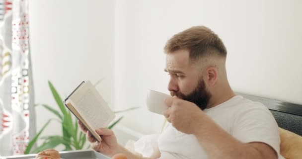 Vista de perto do homem concentrado no meio dos anos 30 lendo livro interessante enquanto toma café da manhã. Bonito cara bebendo chá e virando página enquanto sentado na cama. Conceito de lazer . — Vídeo de Stock